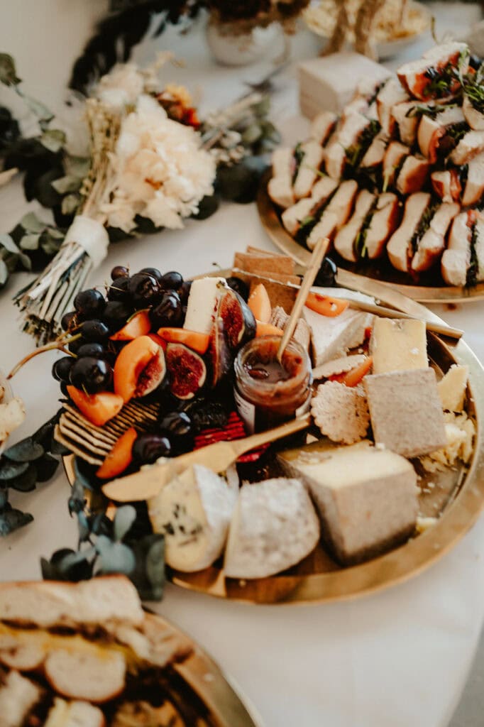 A platter of assorted cheeses, fruits, and jam sits on a table, along with other food items and floral decorations.