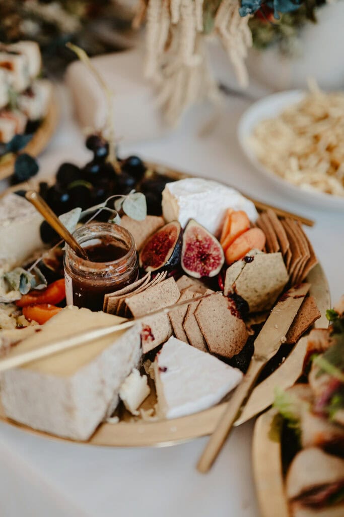 A close-up of a charcuterie board with assorted cheeses, figs, grapes, crackers, and a jar of jam. A knife rests on the side of the board. In the background, there is a bowl of chips.
