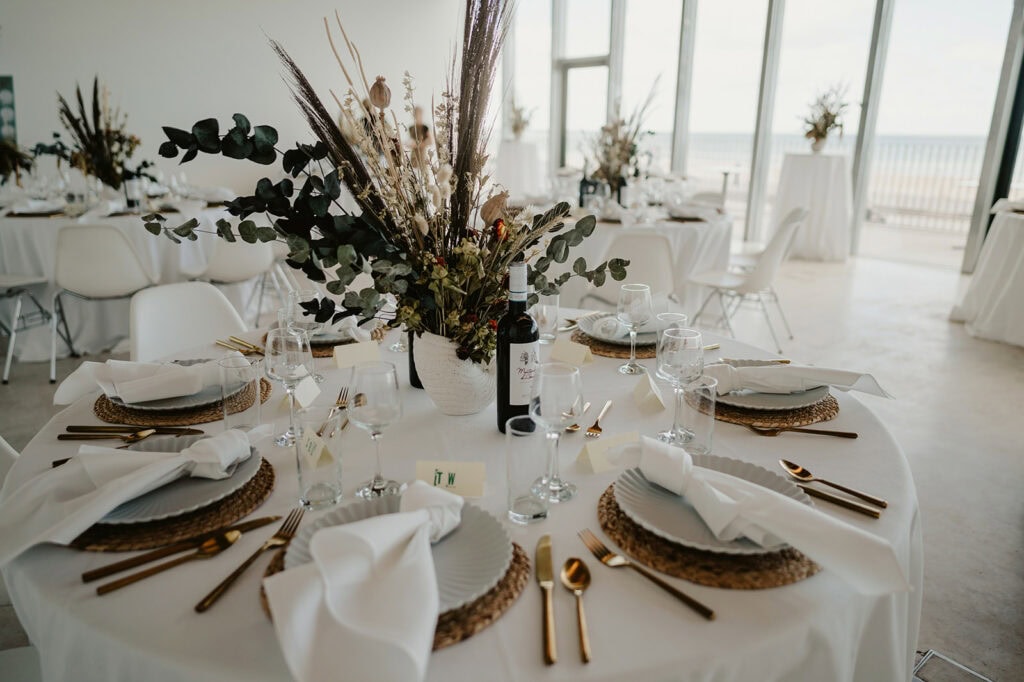 A round table is elegantly set for a meal with white tablecloth, gold utensils, white plates, and a central floral arrangement in a bright room with large windows.