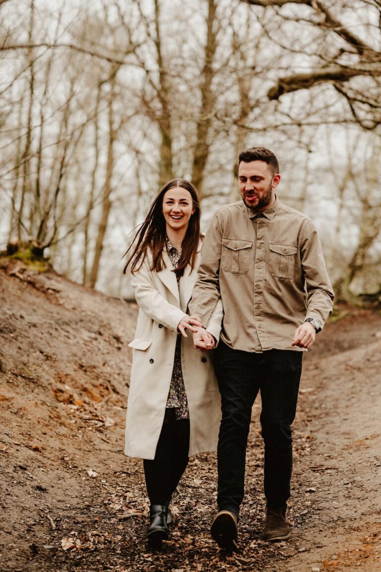 A man and woman walk hand in hand on a muddy path through a wooded area, both smiling and dressed in casual clothes and jackets.