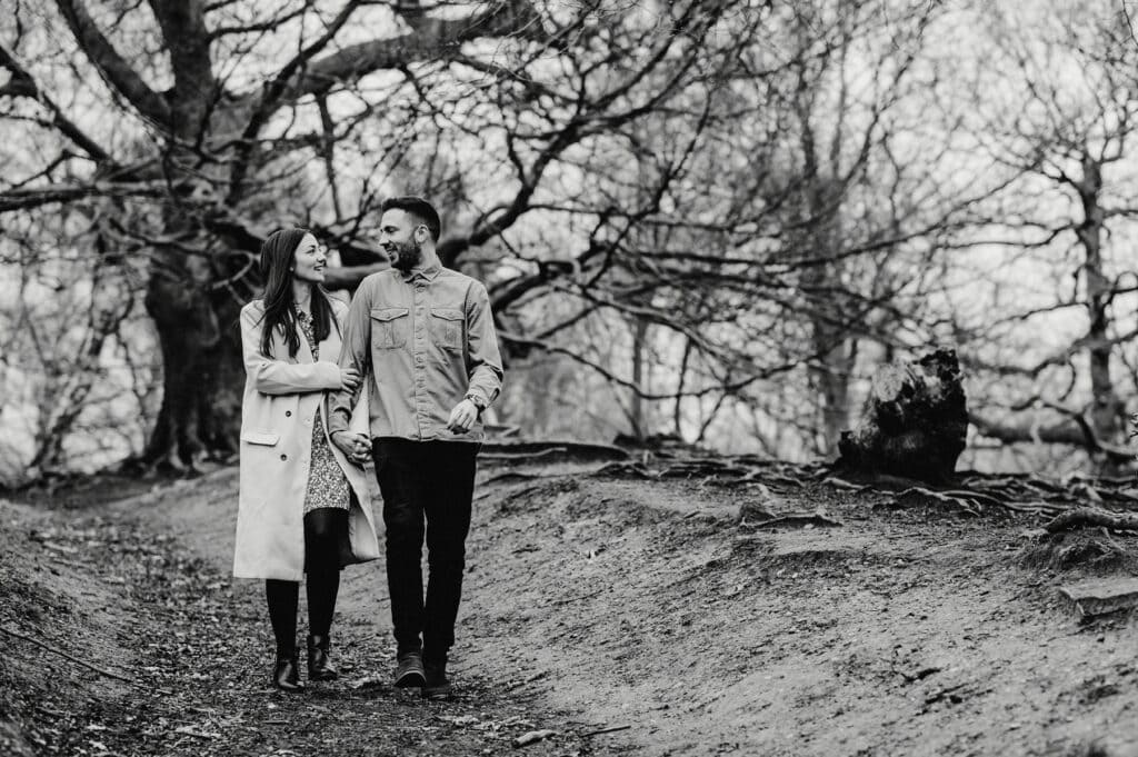 A couple walks hand in hand on a forest path lined with trees. The woman wears a long coat and the man wears a jacket and dark pants. They look at each other affectionately. The scene is in black and white.