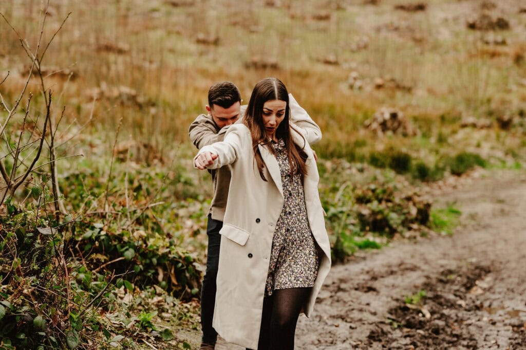 A man and a woman walk down a muddy path in a grassy field. The woman wears a beige coat and floral dress, while the man, slightly behind her, appears to guide her by holding her shoulders.
