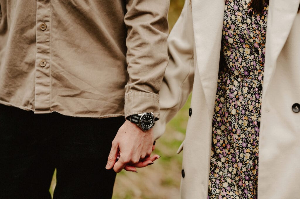 Two people are holding hands. One is wearing a beige shirt and black pants, while the other is wearing a floral dress and white coat.
