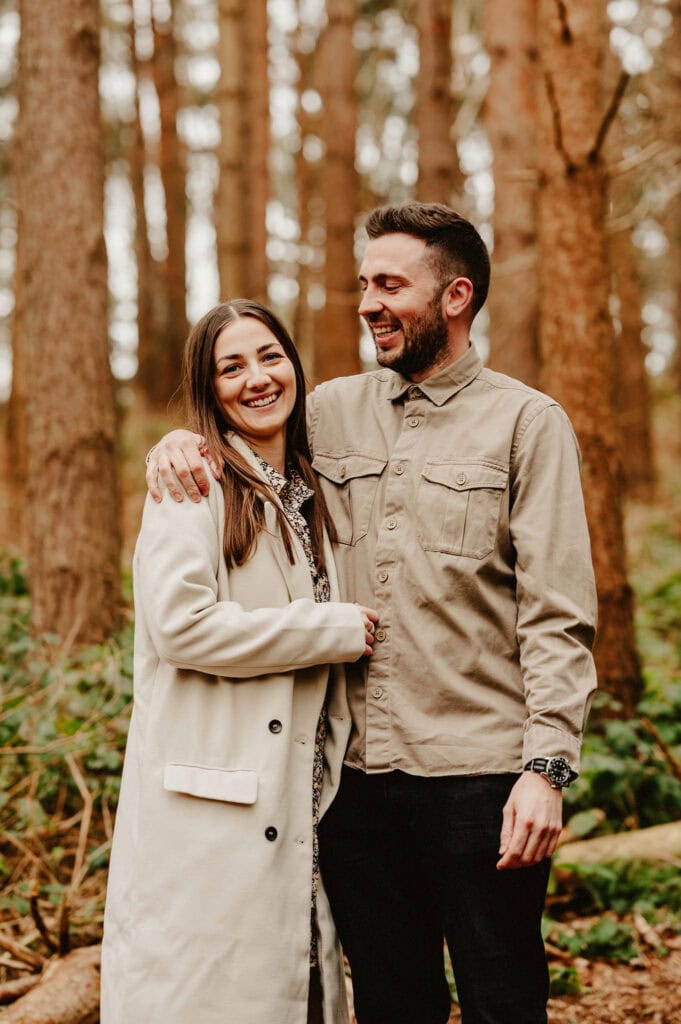 A woman and a man stand close together in a forest. The woman is wearing a light-colored coat, and the man is wearing a beige jacket. Both are smiling.