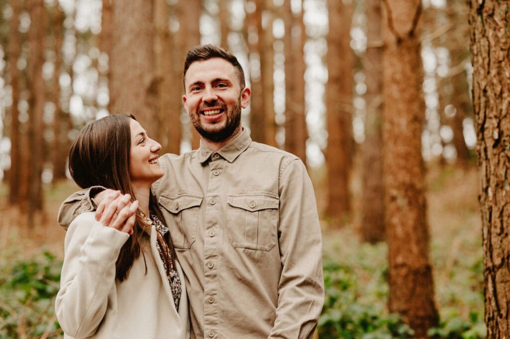 A smiling man and woman stand close together in a forest, with trees and greenery in the background. The man has his arm around the woman, and they both appear happy.