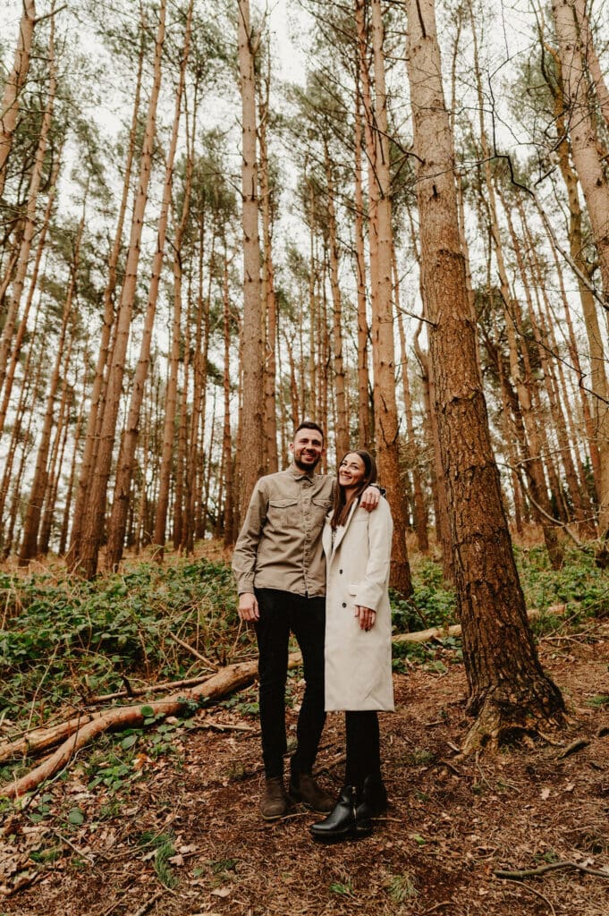 A man and a woman stand side by side in a forest. The man is wearing a khaki jacket and black pants, while the woman is in a light long coat and a beanie. Tall trees fill the background.