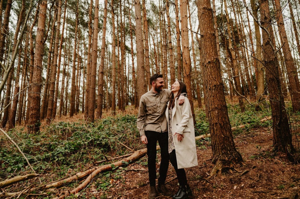 A man and woman stand together in a dense forest. The man wears a brown jacket and the woman wears a long beige coat. They appear to be smiling at each other.