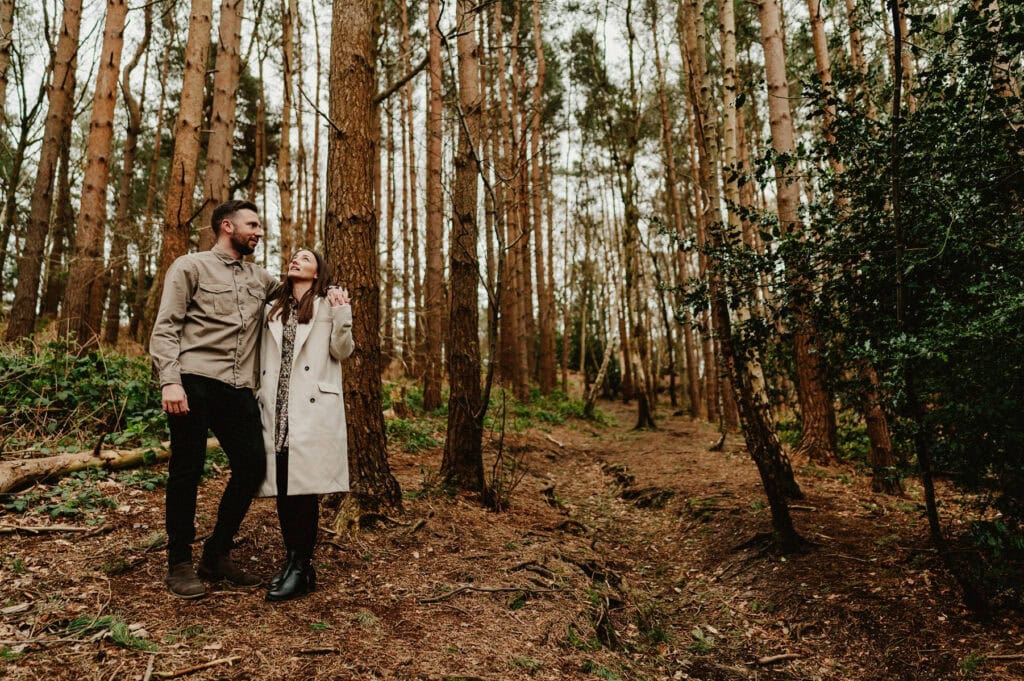 A man and woman are standing in a forest, surrounded by tall trees, wearing casual outdoor clothing. The woman looks up while gently touching the man's chest.