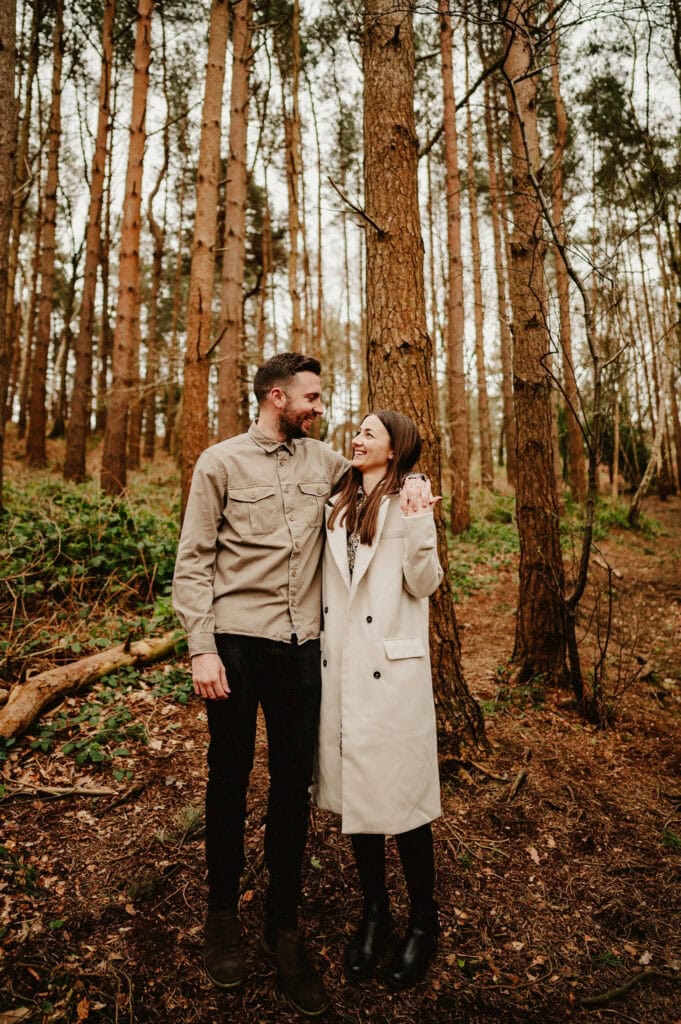 A man and woman stand close together, smiling and looking at each other, in a forest setting with tall trees and scattered leaves on the ground.