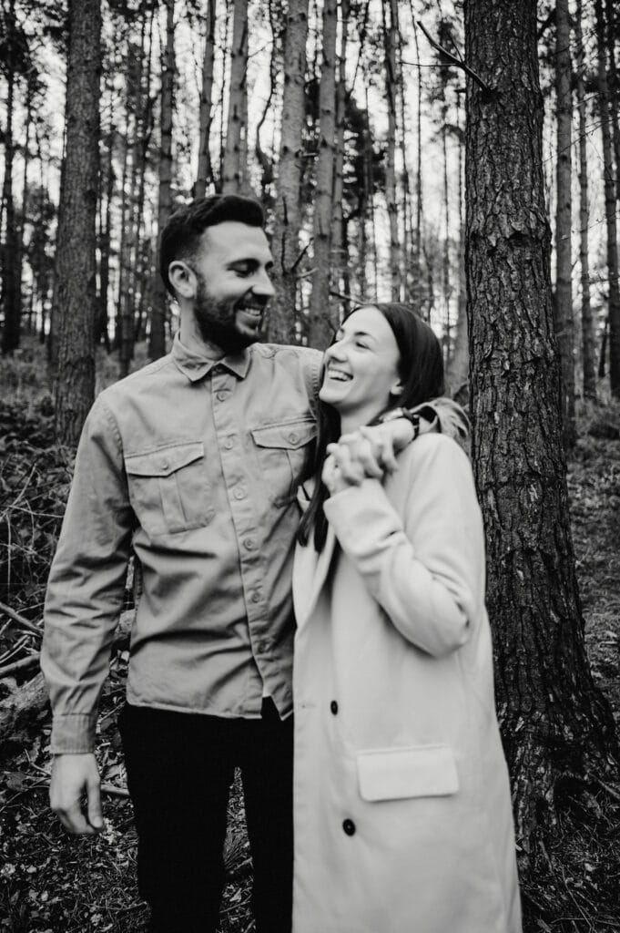 A man and woman smiling and embracing each other while standing in a forest.
