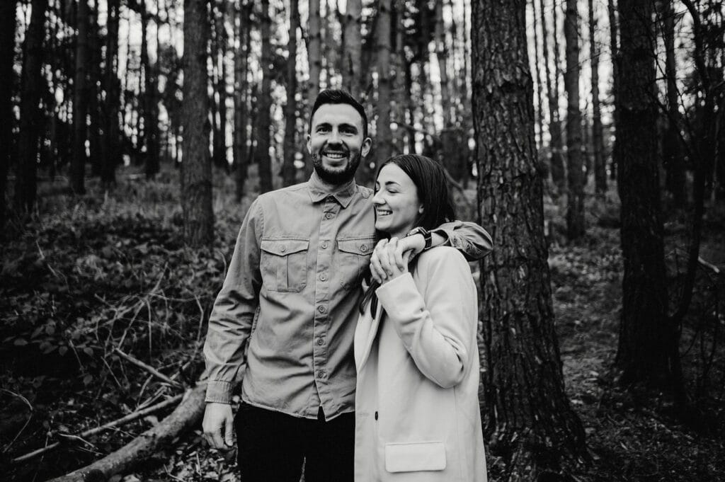 A man and woman stand in a forest, smiling and looking to the side. The woman, wearing a coat, is holding the man's arm. The scene is in black and white.