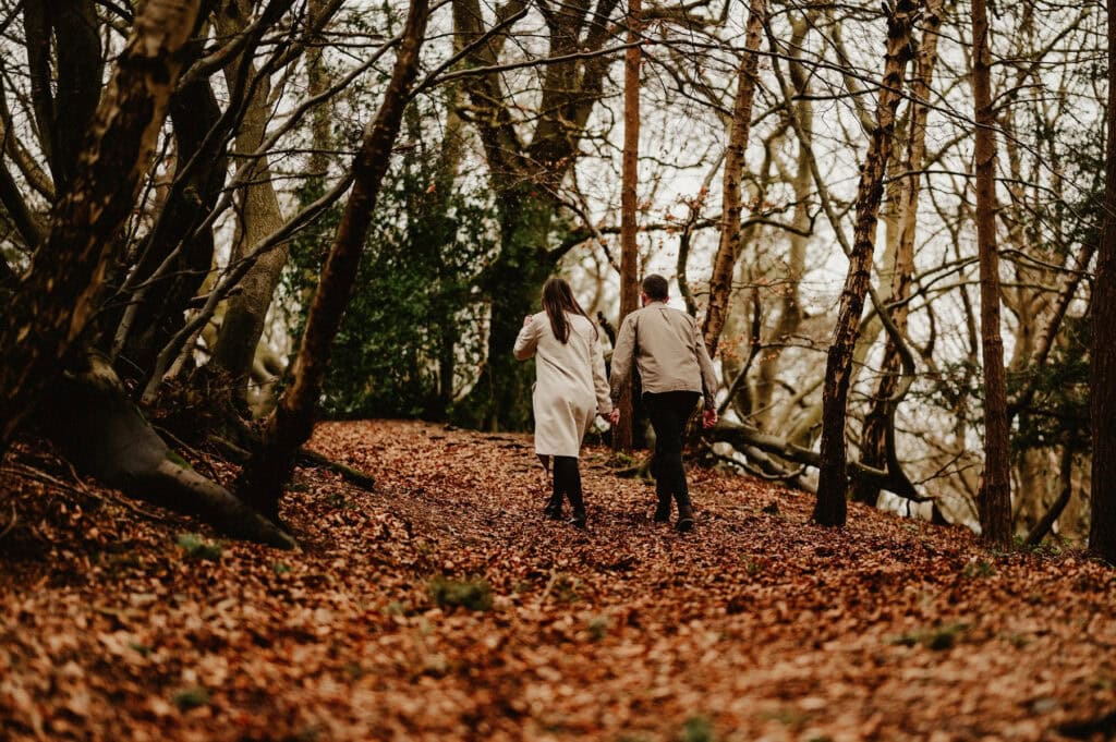 Two people holding hands walk through a forest with fallen leaves on the ground, surrounded by bare trees.