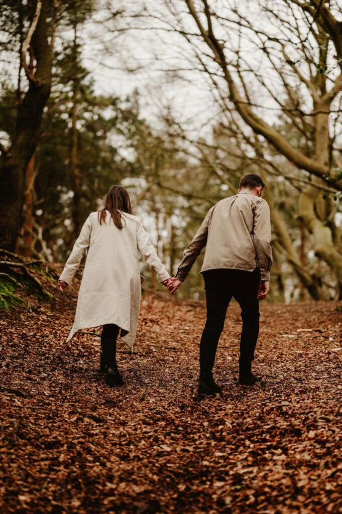 A couple, both wearing coats, walk hand-in-hand up a leaf-covered forest path surrounded by leafless trees.