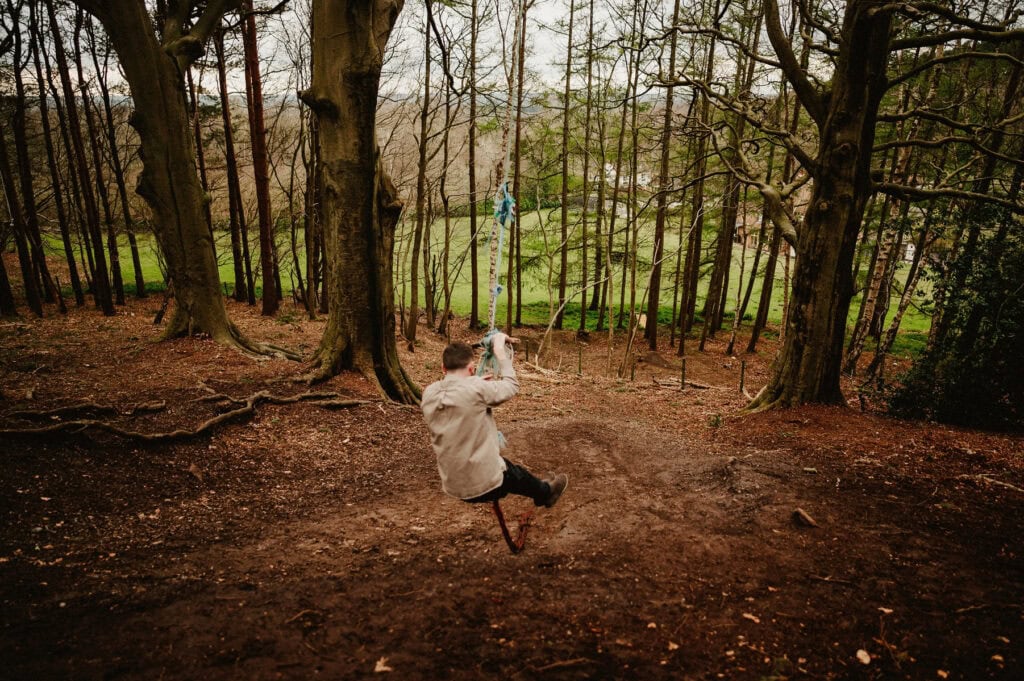 A person swings on a rope swing in a forested area with tall trees and sparse undergrowth. The background shows a grassy field beyond the trees.