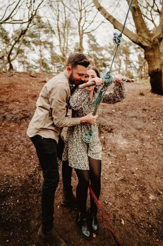 A person in a floral dress holds onto a rope swing with another person in a beige jacket standing behind them, in a wooded area.