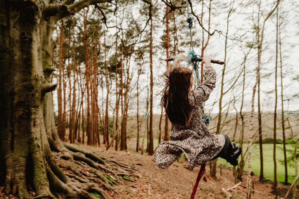 A person with long hair wearing a floral dress swings on a rope swing attached to a large tree in a forested area.
