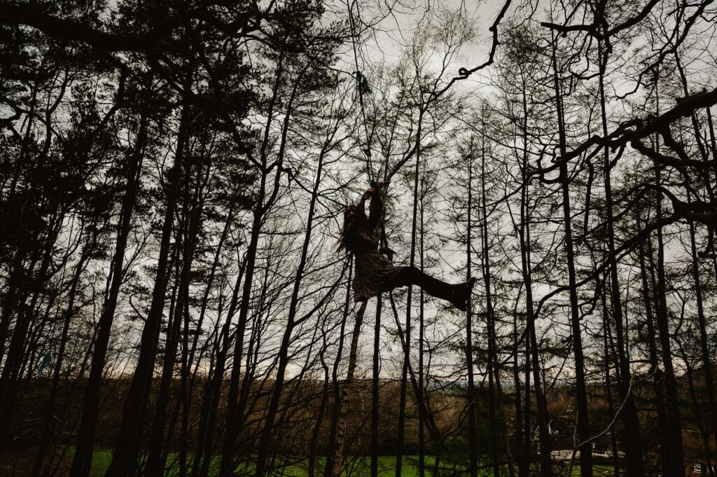 A person swings from a rope in a forest, surrounded by tall trees with bare branches. The sky is overcast, and the ground below is lush and green.