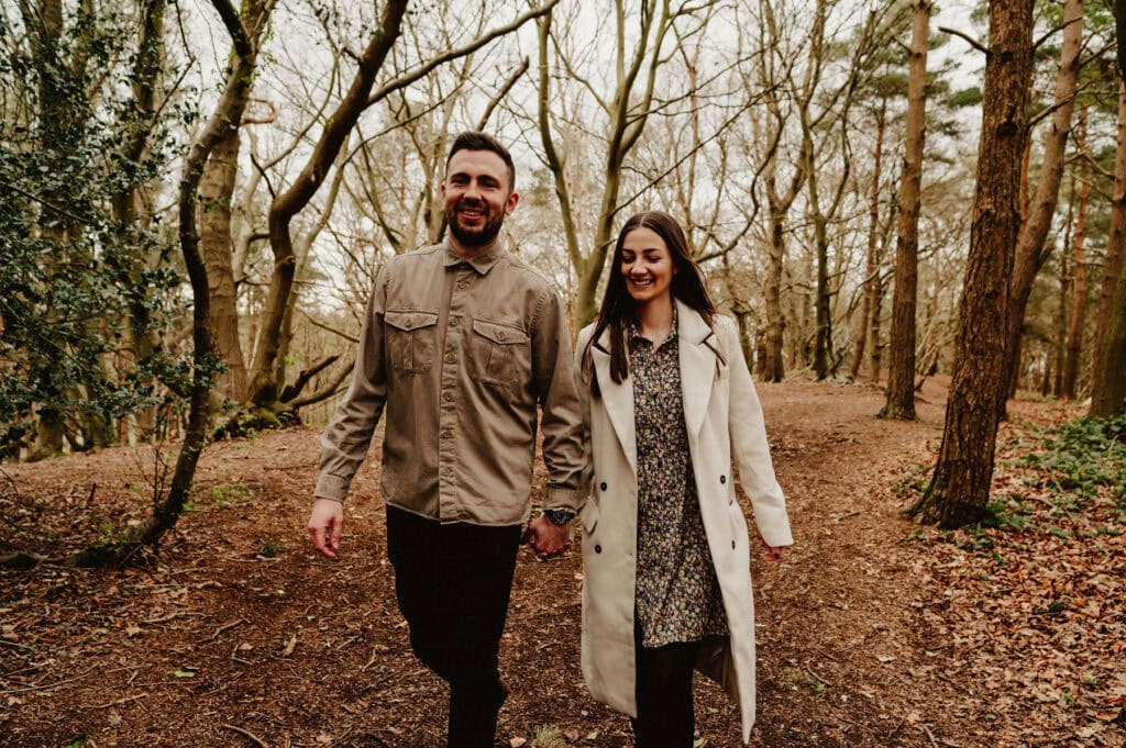 A man and a woman walk hand in hand on a forest path. The man wears a beige shirt and black pants, while the woman wears a light coat over a floral dress. Trees surround them and leaves are on the ground.
