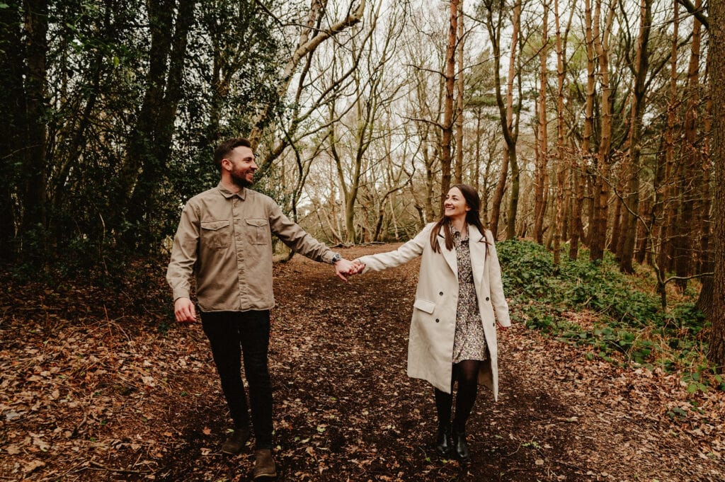 A man and woman holding hands, walking through a forest path covered in leaves, surrounded by trees and greenery.