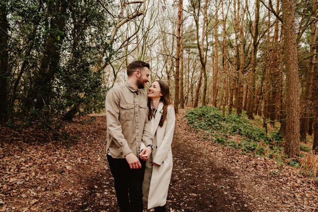 A couple walks hand in hand on a leaf-covered path through a forest, smiling at each other.