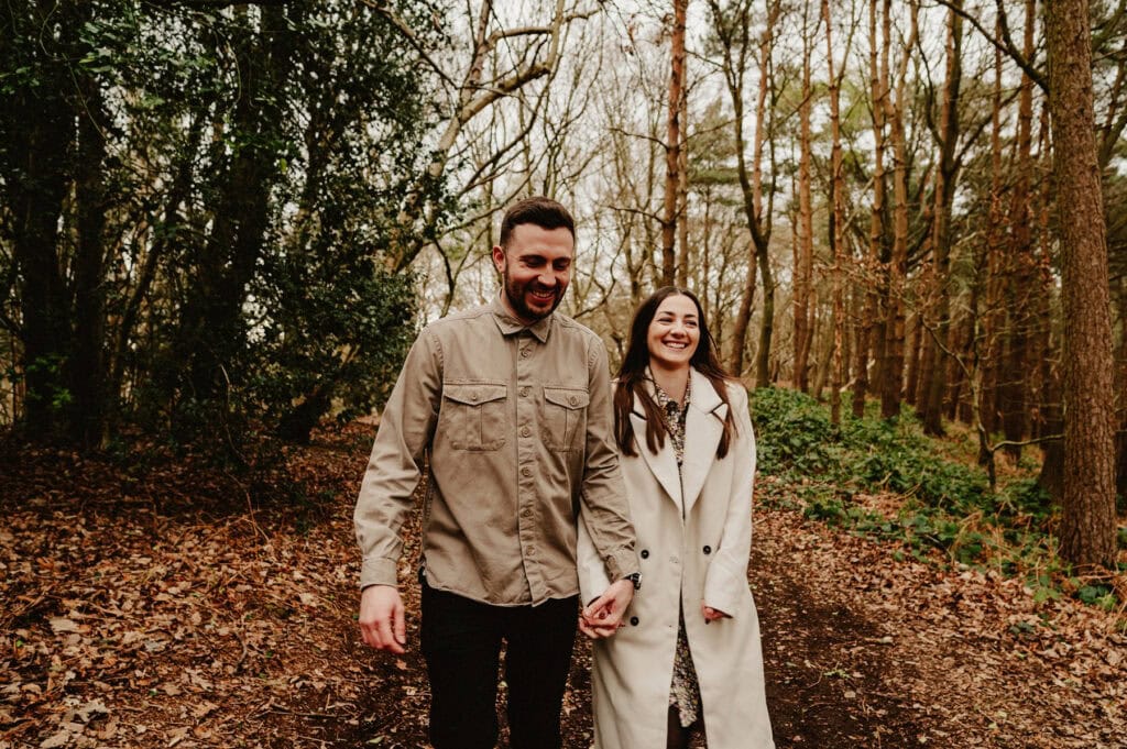 A man and a woman are walking hand in hand through a wooded area on a dirt path, both smiling and wearing casual clothing. The ground is covered in fallen leaves.