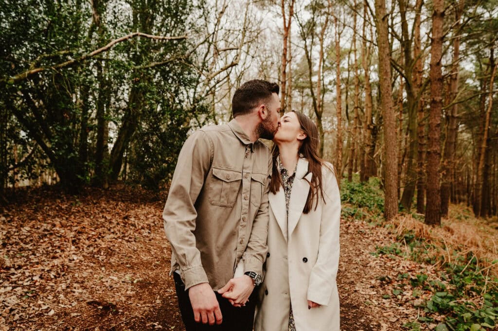 A couple, holding hands and wearing light jackets, kisses while walking on a leaf-strewn path through a dense forest.