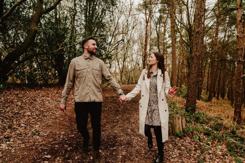 A man and a woman holding hands walk on a forest path surrounded by trees and fallen leaves.