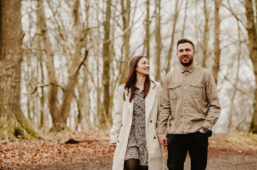 A woman in a light coat and dress and a man in a beige shirt walk hand-in-hand through a forest with bare trees. They both appear content, with the woman looking at the man and the man looking ahead.