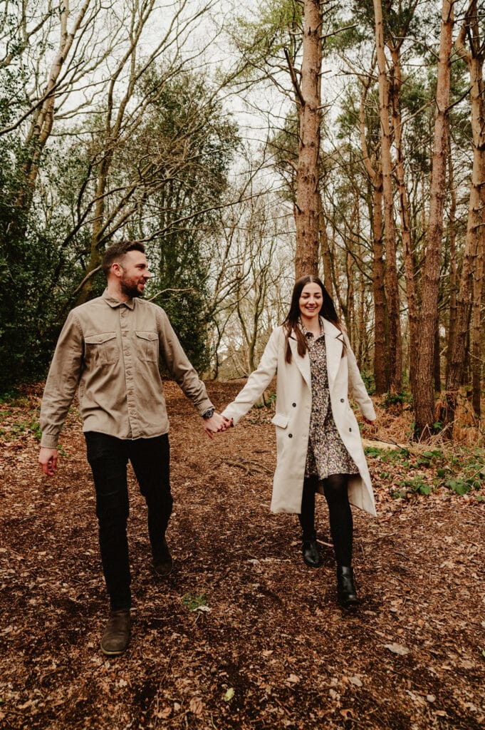 A man and woman hold hands while walking on a forest path covered with fallen leaves, surrounded by tall trees. They are both smiling and dressed in casual attire.
