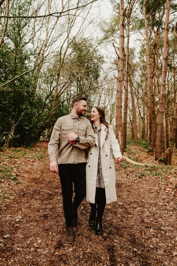 A man and woman walk hand-in-hand through a wooded area with trees and dry leaves on the ground. Both are smiling and wearing coats.