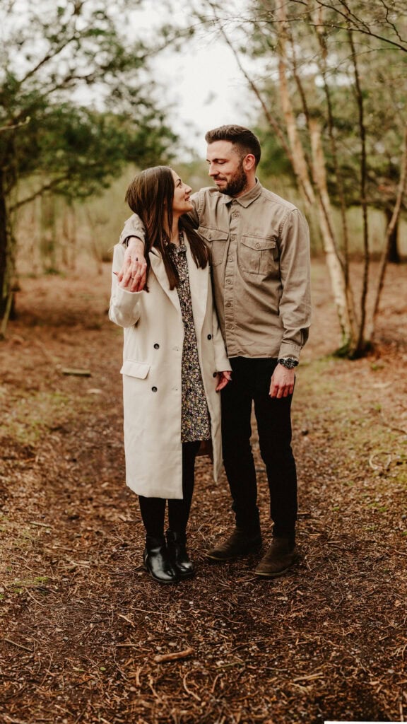 A man and woman stand on a wooded path. The man has one arm around the woman's shoulders. Both are looking at each other, smiling.