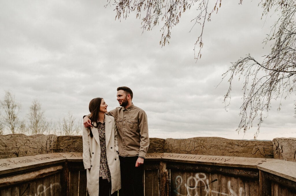 A couple stands together under a cloudy sky, embraced affectionately. They are seen at a wooden lookout spot with graffiti and engraved text on the railing. Sparse trees are in the background.