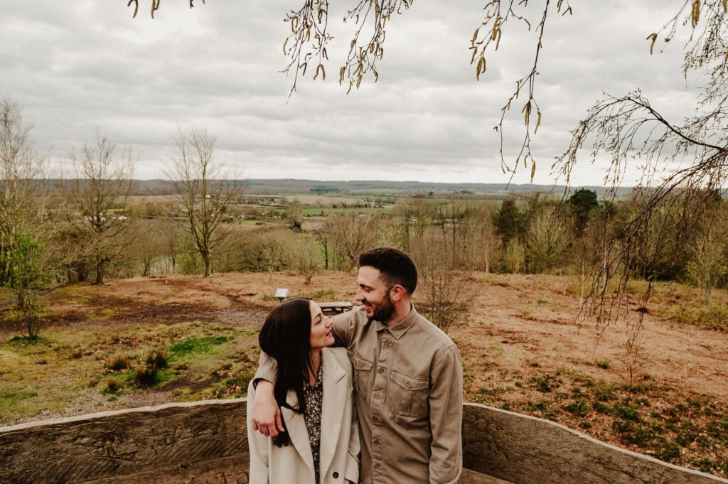A couple stands outside, with the man’s arm around the woman, against a backdrop of an expansive, hilly landscape under a cloudy sky.