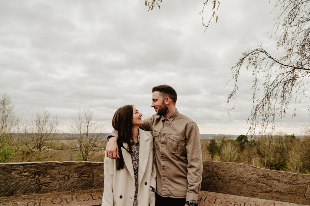A man and a woman stand outdoors with trees and cloudy sky in the background. The man has his arm around the woman's shoulders as they look at each other and smile. They are both wearing jackets.