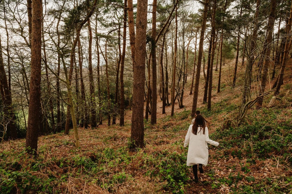 A person in a white coat walks through a dense forest with tall trees and a ground covered in fallen leaves and vegetation.