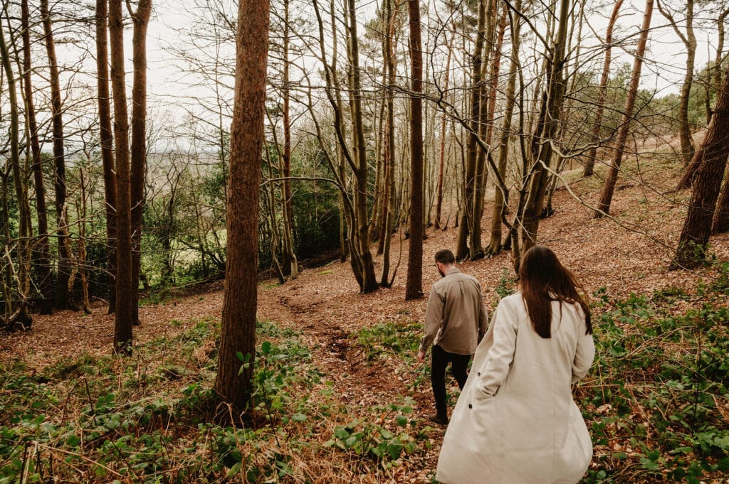 A man and woman walk down a leaf-covered forest path surrounded by tall trees.