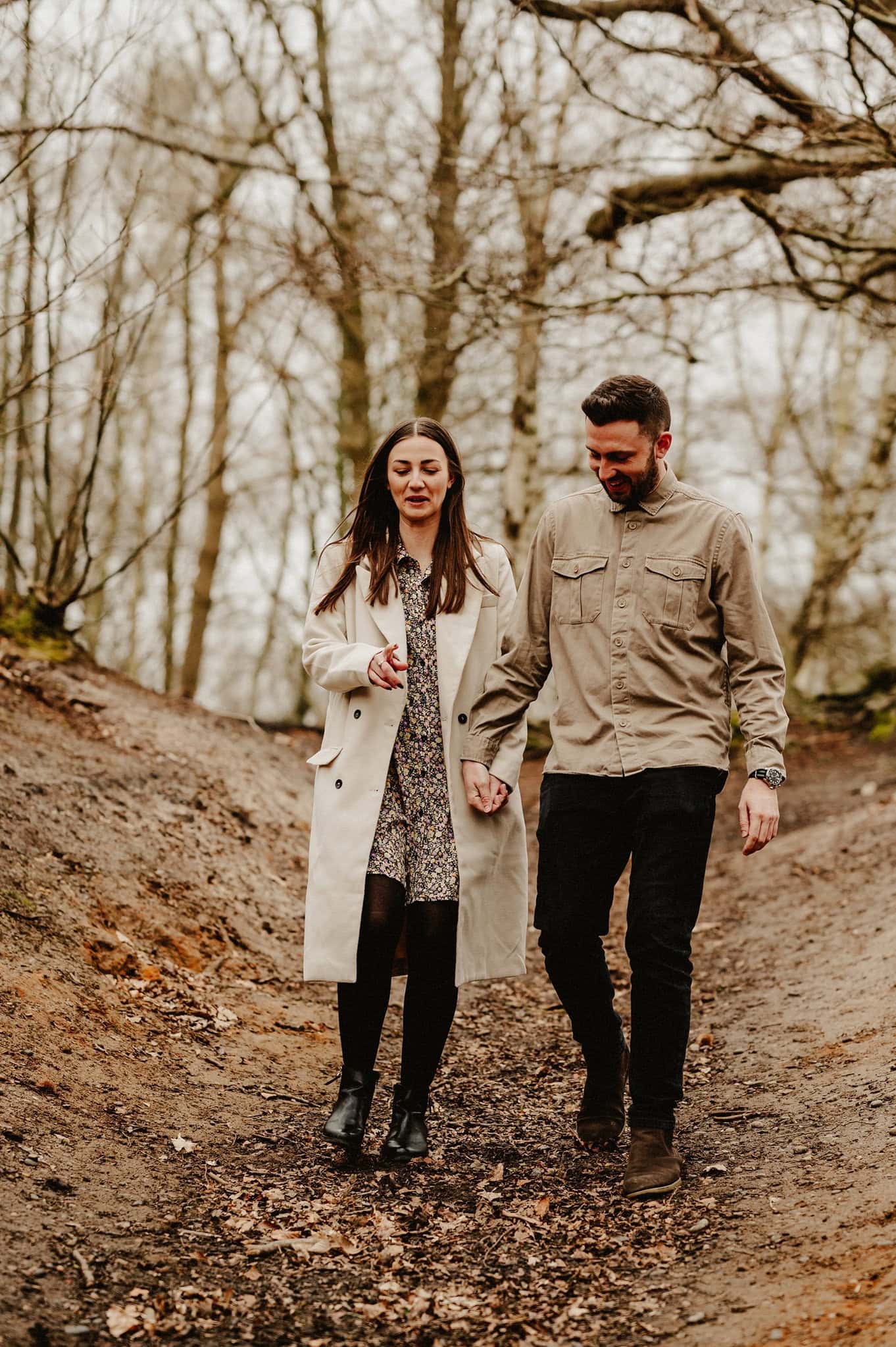 A man and woman holding hands while walking on a dirt path in a wooded area. Both are dressed in casual attire with coats. The background is filled with bare trees.