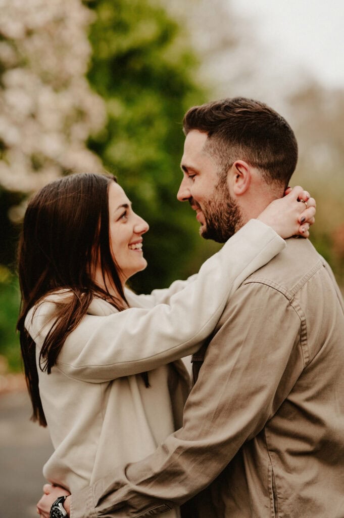 A man and woman stand facing each other outdoors, smiling, with the woman embracing the man around his neck.