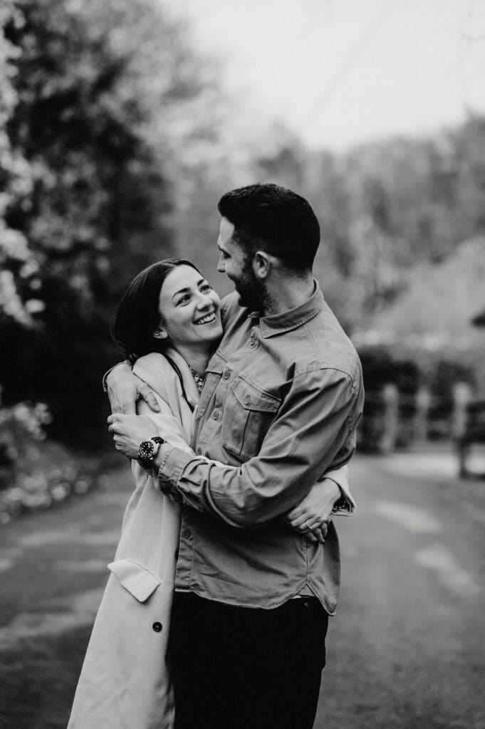 A man and a woman stand embracing on a path in an outdoor setting. The man faces the camera while the woman looks up at him, both smiling. The background is slightly blurred. Black and white image.