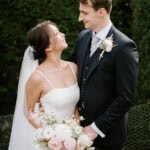 A bride in a white dress and groom in a navy suit stand closely together outdoors, smiling at each other. The bride holds a bouquet of white and pink flowers.