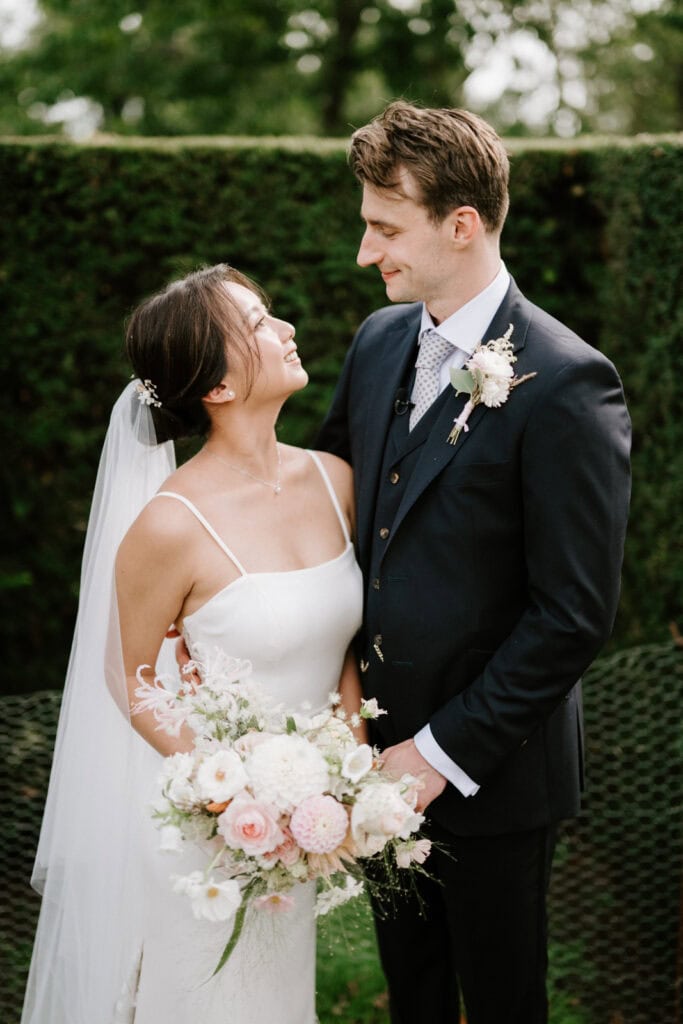 A bride in a white dress and groom in a navy suit stand closely together outdoors, smiling at each other. The bride holds a bouquet of white and pink flowers.