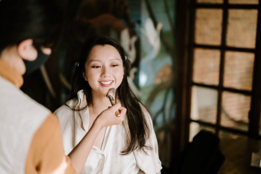 A woman is smiling while another person, partially visible, applies makeup to her face with a brush inside a room with a mural and a window.