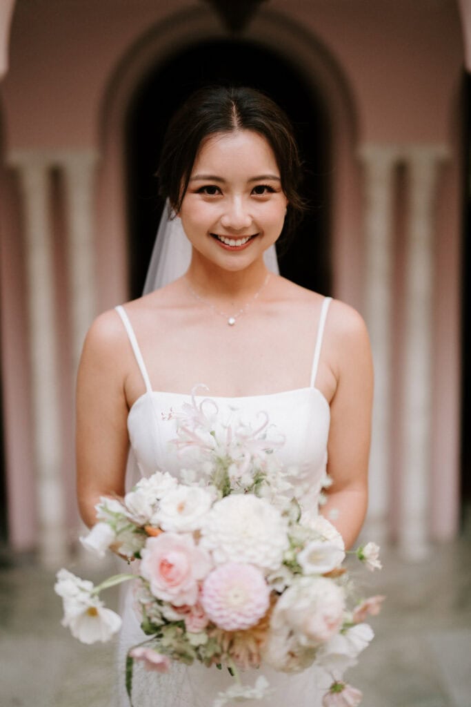A woman in a white wedding dress holds a bouquet of flowers, standing in front of a building with arched doorways.