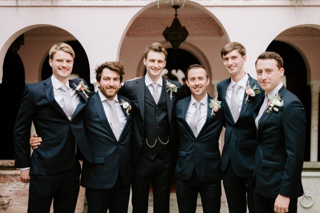 Six men in suits stand together under an archway. Five wear matching navy blue suits with blue ties and boutonnieres, while the man in the center wears a similar suit with a vest.