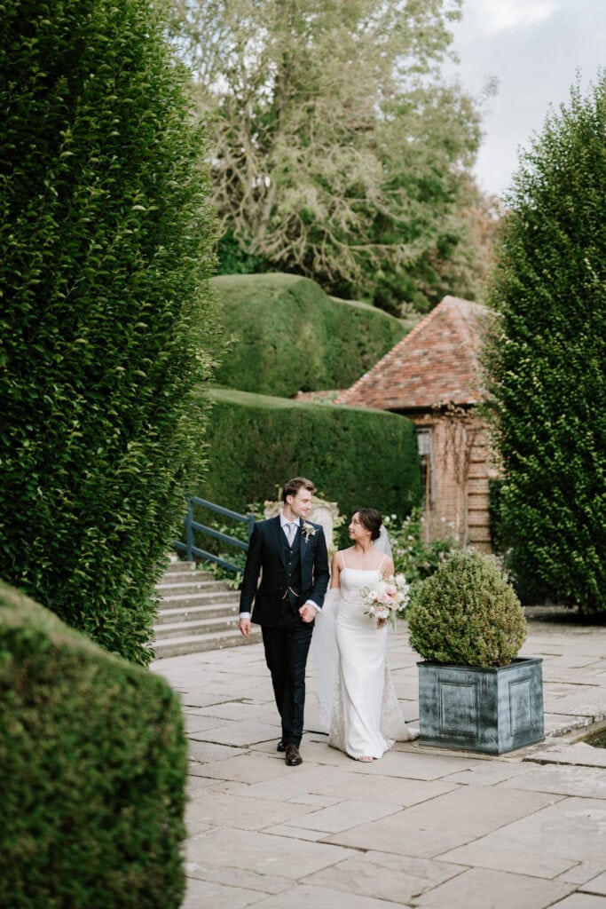 A newlywed couple walks down a stone path surrounded by manicured hedges and greenery; the groom wears a black suit and the bride wears a white dress while holding a bouquet of flowers.