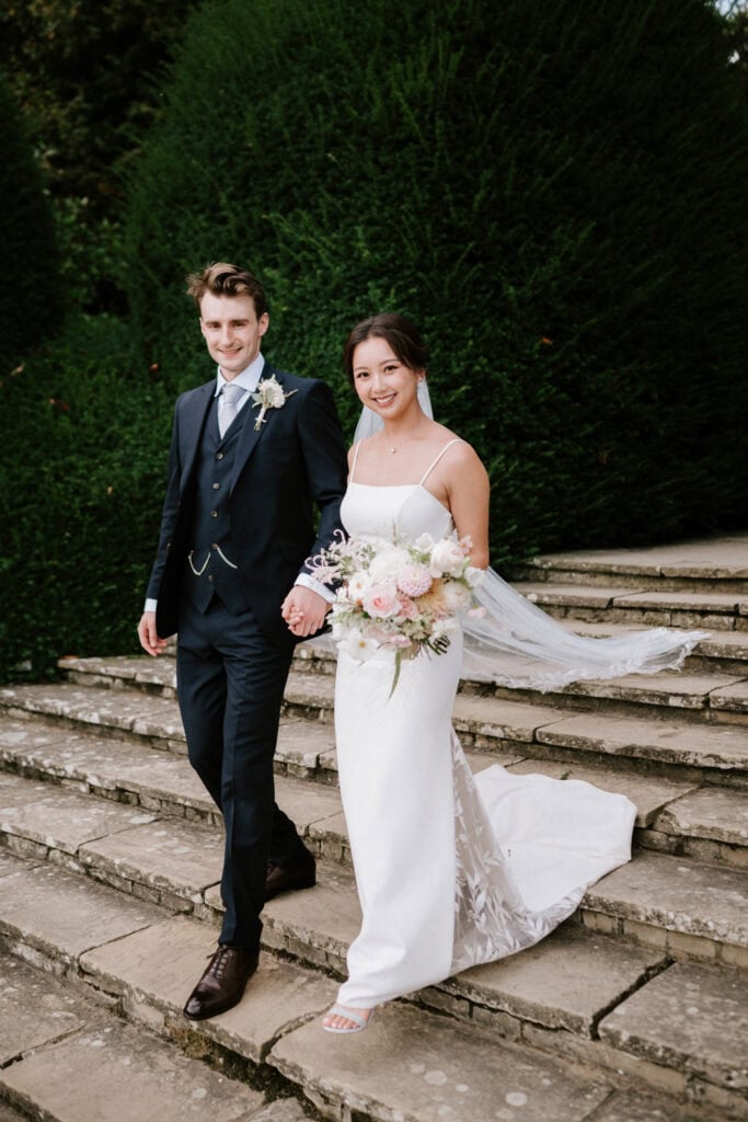 A bride in a white gown and a groom in a dark suit walk hand in hand down stone stairs, smiling. The bride holds a bouquet of white and pink flowers.