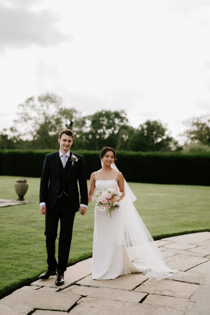 A bride and groom walk hand in hand in an outdoor garden setting, with the bride holding a bouquet of flowers and both dressed in formal wedding attire.