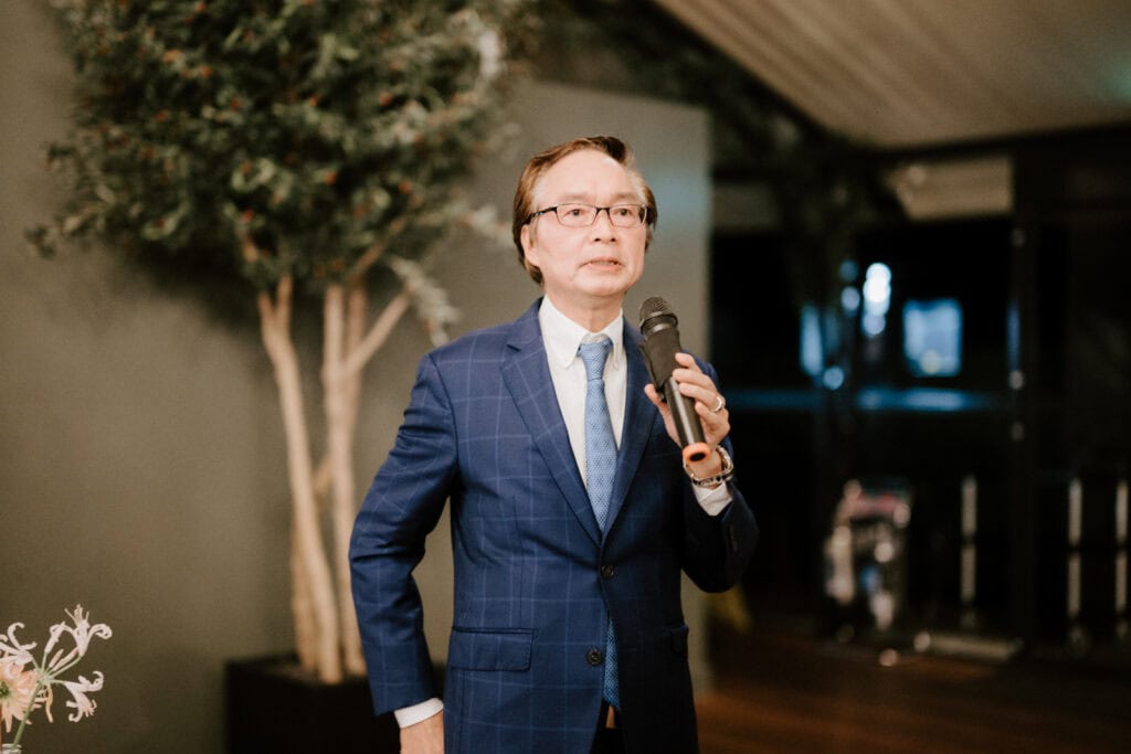A man in a blue suit speaking into a microphone while standing indoors near a plant.