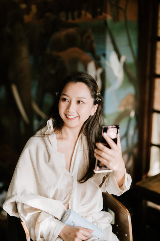 A woman with long hair sits in a chair holding a phone and smiling against a backdrop with animal murals. She wears a light-colored shirt and hair clips.