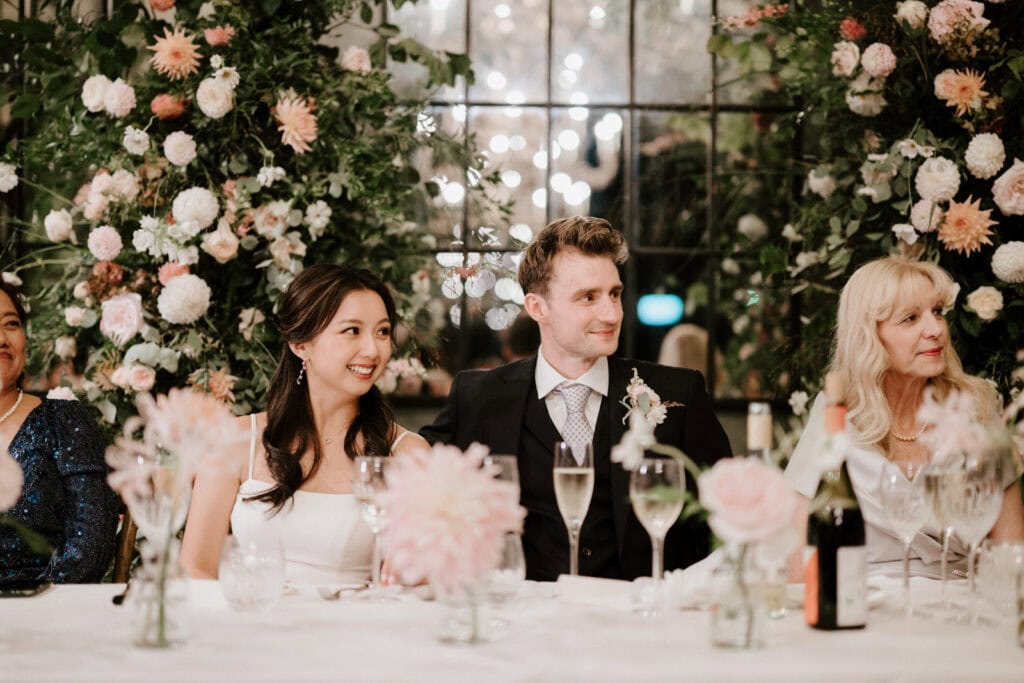A bride and groom sit at a decorated table, surrounded by flowers and guests during their wedding reception. The couple is smiling and looking towards someone off-camera.